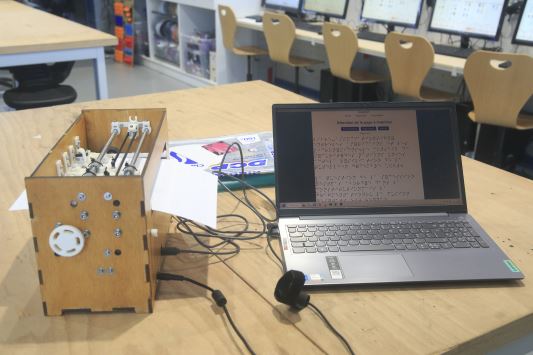 A varnished wooden BrailleRAP sits on a workbench. Next to it, a laptop with AccessBrailleRAP software displays a Braille page.